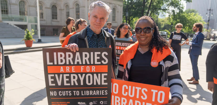 Man-And-Woman-Holding-Signs-At-Rally-About-Library-Budget-Cuts